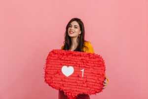 Teenager girl in bright outfit is smiling, holding red "Like" sign from Instagram on pink backgroun